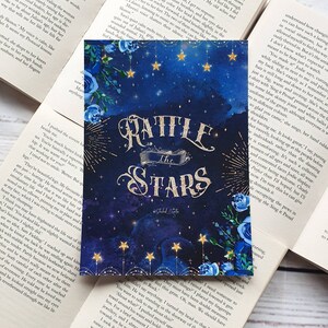 May include: A book cover with a blue watercolor background featuring stars and blue flowers. The title "Rattle the Stars" is written in gold glitter lettering.