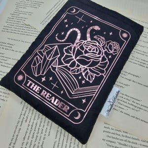 以下が含まれることがあります： 黒いタロットカードポーチ。バラ、ヘビ、クリスタル、そして「THE READER」の文字が描かれたローズゴールドのデザイン。ポーチは柔らかい濃い色の生地で作られており、「Needles Riddles」と書かれた小さなタグが付いています。