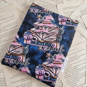 Può includere: Una custodia per libri in tessuto blu e rosa con un design di una strega e libri. Il testo sulla custodia recita "Bad Witch Book Club".