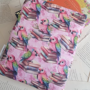 May include: A pink and white fabric with a repeating pattern of colourful parrots perched on stacks of books. The parrots have green, blue, and yellow feathers. The books are brown and have gold lettering. The background is a light pink with a subtle texture.