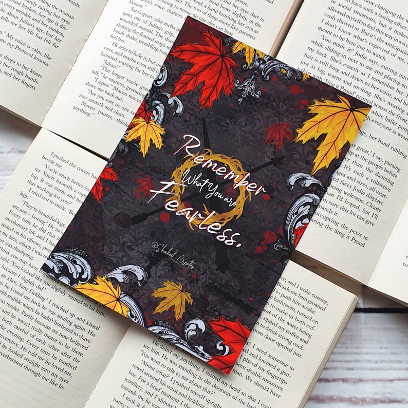 May include: A black and white bookmark featuring a wreath of autumn leaves and the text "Remember What You Are Fearless." The text is in a white circle with a black outline. The background is a dark grey with a floral pattern.