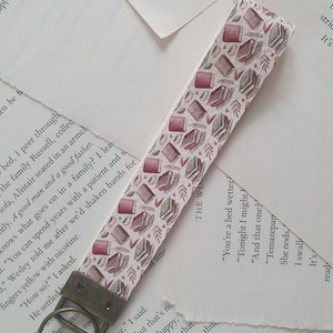 May include: A white and pink fabric bookmark with a repeating pattern of stacked books and leaves. The bookmark has a brass key ring attached.