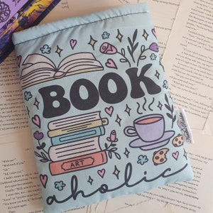 Può includere: Una borsa tote blu chiaro a tema libro con il testo "BOOKaholic" in lettere nere. La borsa presenta una pila di libri, una tazza di caffè e un biscotto. I libri hanno la parola "ART" sulla copertina.