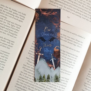 May include: A blue bookmark with a dark blue background and a white mountain scene. The text "Our Stories are Worth Telling" is written in white with a dark blue border. Two swords are visible in the foreground.