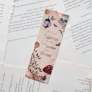 May include: A bookmark with a beige background and a quote that reads "If I cannot be better, then I will become much worse". The bookmark features a skull, a lantern, mushrooms, and acorns in a whimsical style.