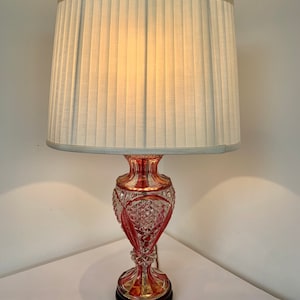 Puede incluir: Una lámpara de mesa de vidrio rosa y transparente con una pantalla plisada blanca. La base de la lámpara es un diseño de vidrio decorativo, facetado y texturizado.