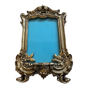 May include: A gold-toned ornate picture frame with a blue background. The frame has a decorative design with intricate details.
