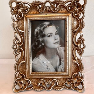 Puede incluir: Un marco de fotos plateado adornado con una abertura de 10,16 cm x 15,24 cm. El marco tiene un diseño detallado e intrincado con una superficie texturizada. Una foto en blanco y negro de una mujer está dentro del marco.