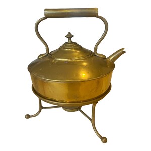 May include: A vintage brass kettle with a stand. The kettle has a round body with a curved handle and a spout. The stand has three legs.