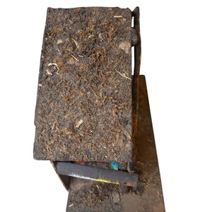 Pode incluir: Uma pequena estrutura de madeira r&uacute;stica com um telhado coberto de musgo. O edif&iacute;cio tem uma apar&ecirc;ncia escura e desgastada, com gr&atilde;o de madeira vis&iacute;vel e uma pequena base angular. O design sugere um artigo artesanal, feito &agrave; m&atilde;o.