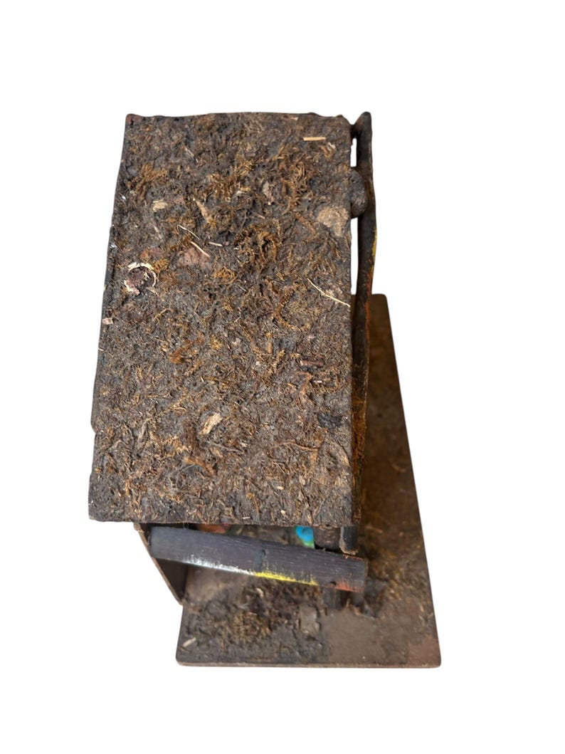 Pode incluir: Uma pequena estrutura de madeira r&uacute;stica com um telhado coberto de musgo. O edif&iacute;cio tem uma apar&ecirc;ncia escura e desgastada, com gr&atilde;o de madeira vis&iacute;vel e uma pequena base angular. O design sugere um artigo artesanal, feito &agrave; m&atilde;o.