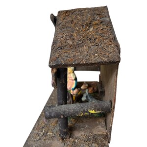 Pode incluir: Um pres&eacute;pio de madeira em miniatura r&uacute;stico com um telhado coberto de musgo. A estrutura apresenta uma moldura castanha escura, um aspeto envelhecido e figuras vis&iacute;veis no interior. A cena est&aacute; sobre um fundo branco.
