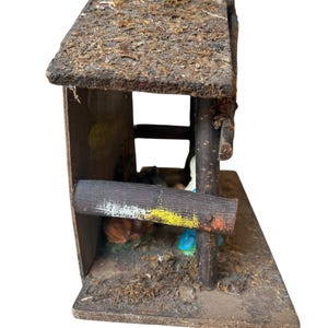 Può includere: Un presepe in miniatura rustico. La struttura in legno presenta un tetto ricoperto di muschio, una base usurata e una staccionata dipinta. All'interno, le statuine raffigurano una scena religiosa con un animale che dorme e figure in abiti blu e bianchi.