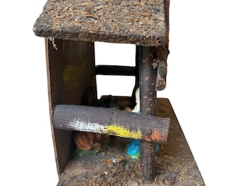 Pode incluir: Um presépio em miniatura rústico. A estrutura de madeira apresenta um telhado coberto de musgo, uma base desgastada e uma cerca pintada. No interior, as figuras retratam uma cena religiosa com um animal a dormir e figuras com vestes azuis e brancas.