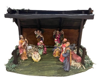 Pode incluir: Um presépio tradicional em um estábulo de madeira com telhado marrom. As figuras incluem Maria, José, o menino Jesus, os três reis magos, um anjo e animais de fazenda. A cena é decorada com palha e vegetação.