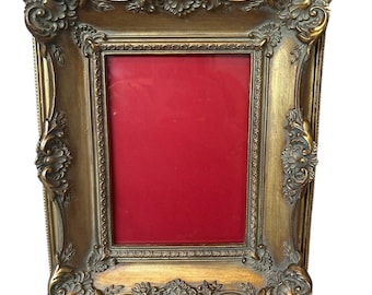 Puede incluir: Marco de fotos dorado ornamentado con interior rojo. El marco presenta detalles de volutas y acentos decorativos en las esquinas. El marco rectangular está diseñado para una fotografía o una obra de arte.