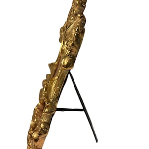 Puede incluir: Marco de fotos dorado y ornamentado con un diseño decorativo y texturizado. El marco está sostenido por un caballete de metal negro, lo que le permite mantenerse en posición vertical. El diseño del marco presenta un patrón detallado y esculpido.