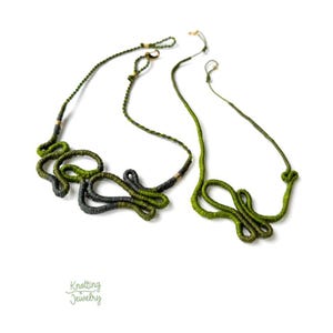 Op de afbeelding: Twee handgemaakte kettingen met een geknoopt ontwerp. De kettingen zijn gemaakt van groen en grijs touwachtig materiaal. De ene ketting heeft een lusontwerp en de andere een meer lineair ontwerp. De kettingen staan op een witte achtergrond.