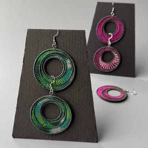 Op de afbeelding: Handgemaakte oorringen in groen, blauw en wit, tentoongesteld op een zwarte standaard. Een ander paar in roze en een enkele roze oorbel zijn ook afgebeeld. De oorbellen hebben een gelaagd, geweven ontwerp en zilverkleurige haken.