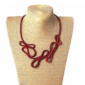 Op de afbeelding: Een rode, abstracte touwketting op een mannequin. De ketting is gemaakt van dik, gedraaid touw en heeft een unieke, organische vorm.
