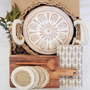 May include: A gift basket with a white ceramic plate with a floral design, a wooden cutting board with a heart-shaped spoon, and a set of woven coasters. The basket is lined with brown wicker.