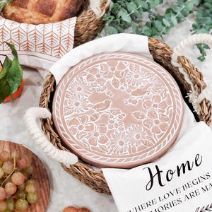 May include: A round terracotta-colored ceramic lid featuring a white embossed floral and bird pattern, nestled inside a woven wicker basket with rope handles. A white cloth with the text Home WHERE LOVE BEGINS AND MEMORIES ARE MADE is draped.