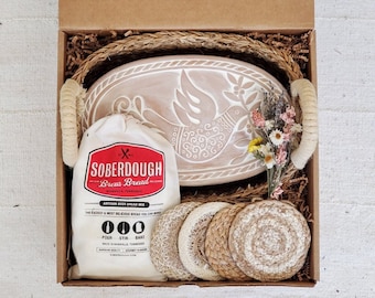 May include: A gift box filled with a white ceramic bread bowl with a dove design, a bag of sourdough bread mix, and four natural woven coasters. The box is lined with brown straw.