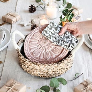 May include: A festive table setting with white plates, gold cutlery, and linen napkins. A woven basket holds a pink, floral-patterned serving dish. The table is decorated with candles, pine cones, and wrapped gifts, creating a warm, inviting atmosphere.