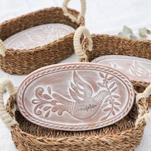 May include: An oval terracotta tray with a white embossed dove carrying an olive branch and the text Miller EST 2023, set inside a woven seagrass basket with thick cream-colored rope handles. The tray features a decorative rope-like border.