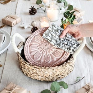 May include: A round, woven seagrass basket with white rope handles containing a dusty pink ceramic plate with a floral relief pattern. A folded white napkin with a black geometric tree print is placed on top of the plate within the basket.