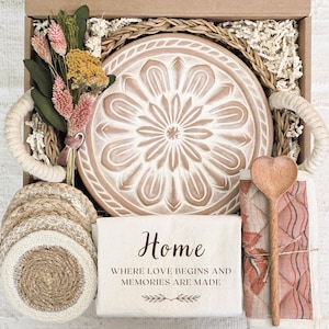 May include: A gift box filled with home decor items, including a wooden serving tray with a floral design, a set of woven coasters, a heart-shaped wooden spoon, and a white tea towel with the text "Home Where Love Begins and Memories Are Made".