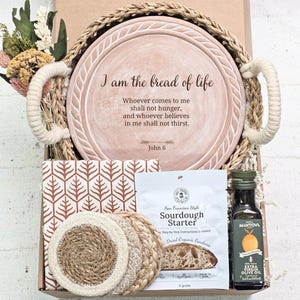 May include: A gift basket with a decorative plate inscribed with "I am the bread of life." The basket includes a sourdough starter, olive oil, coasters, and a patterned fabric. Dried flowers and woven handles complete the presentation.