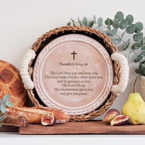 May include: A round wooden serving tray with rope handles. The tray has a white background with a brown border and a cross at the top. The text "Numbers 6:24-26" is written in brown below the cross. The text below reads "The Lord bless you and keep you; The Lord make His face shine upon you, and be gracious to you; The Lord lift up His countenance upon you, and give you peace."