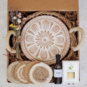 May include: A gift box containing a round terracotta-colored plate with a white floral pattern, a woven tray with handles, a small bottle of dark liquid, a white box labeled Jocelyn & Co with olive oil and sea salt crackers, and dried flowers.