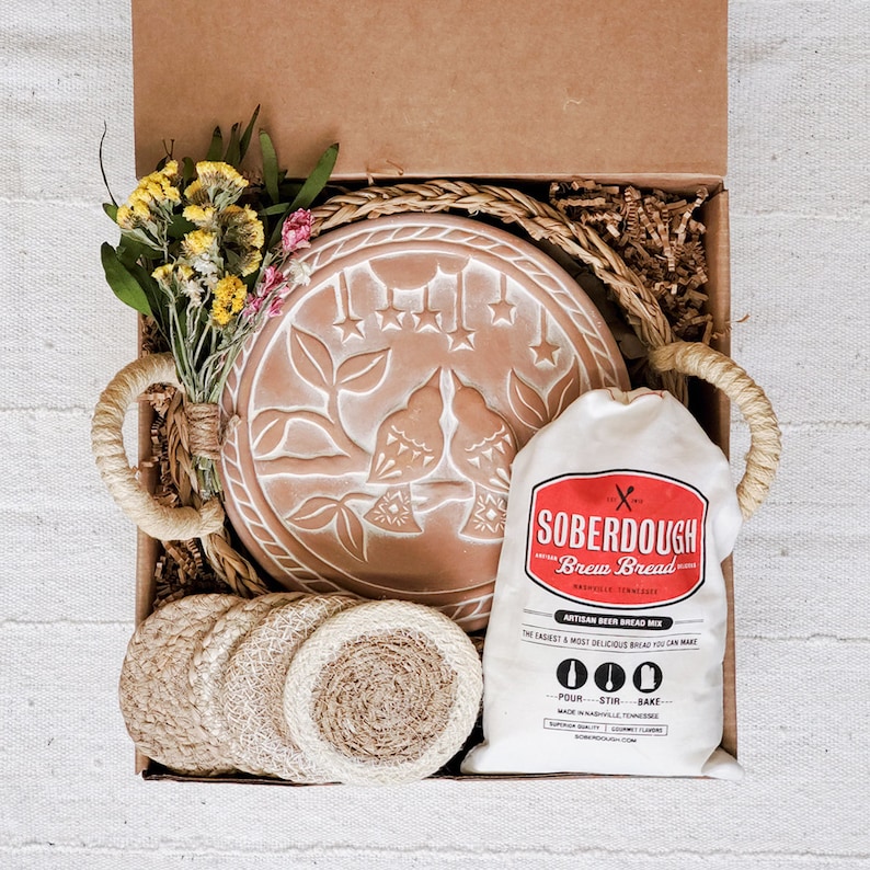 Puede incluir: Una caja de regalo llena de un plato para hornear pan de terracota con un dise&ntilde;o de p&aacute;jaro, una bolsa de mezcla para pan de masa madre y posavasos tejidos. La bolsa tiene el texto "SOBERDOUGH Brew Bread" e instrucciones para "POUR - STIR - BAKE".