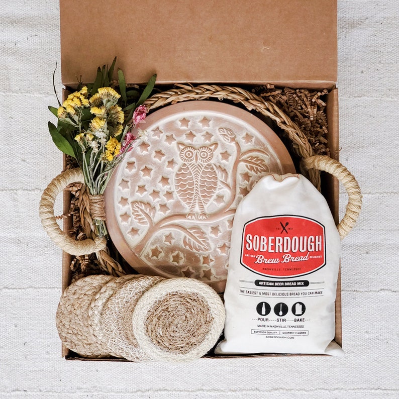 Puede incluir: Una caja de regalo llena de una fuente para hornear de cer&aacute;mica marr&oacute;n con un dise&ntilde;o de estrellas y b&uacute;hos, una bolsa de mezcla para pan de masa madre y posavasos de fibra natural. La bolsa de mezcla tiene el texto "SOBERDOUGH Brew Bread Artisan Beer Bread Mix" impreso en ella.