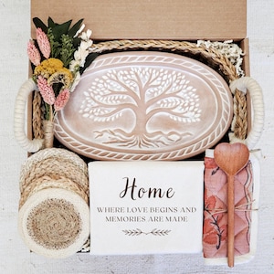 May include: A gift box filled with home decor items. Includes a decorative oval platter with a tree design, coasters, a wooden spoon, a tea towel, and a bouquet of dried flowers. The box has the text "Home" on the front.