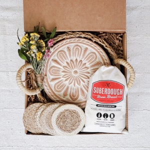 Puede incluir: Una caja de regalo llena de suministros para hornear, que incluyen una mezcla de pan de masa madre, una campana de pan de cer&aacute;mica y posavasos de fibra natural. La caja est&aacute; forrada con papel marr&oacute;n y paja.