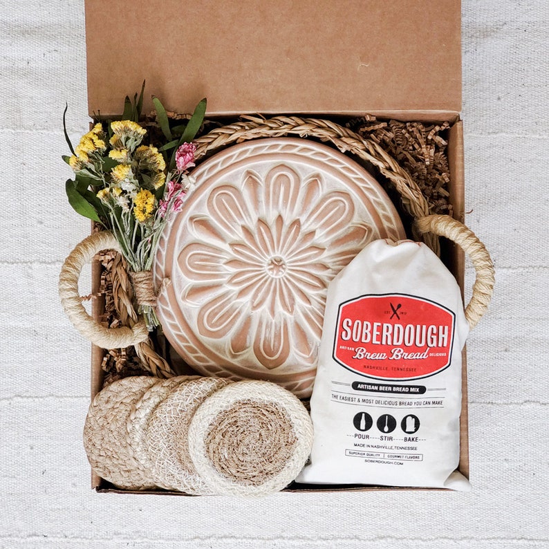 Puede incluir: Una caja de regalo llena de suministros para hornear, que incluyen una mezcla de pan de masa madre, una campana de pan de cer&aacute;mica y posavasos de fibra natural. La caja est&aacute; forrada con papel marr&oacute;n y paja.