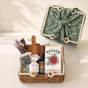May include: A woven basket containing a wooden cutting board, a small bag of Fleur de Sel, a bottle of Mantova extra virgin olive oil, a box of Hayden Flour Mills artisan bread flour, a patterned cloth, and dried flowers, with a matching fabric lid.