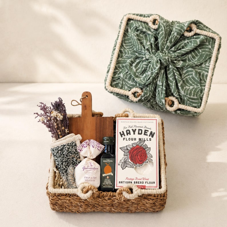 May include: A woven basket containing a wooden cutting board, a small bag of Fleur de Sel, a bottle of Mantova extra virgin olive oil, a box of Hayden Flour Mills artisan bread flour, a patterned cloth, and dried flowers, with a matching fabric lid.