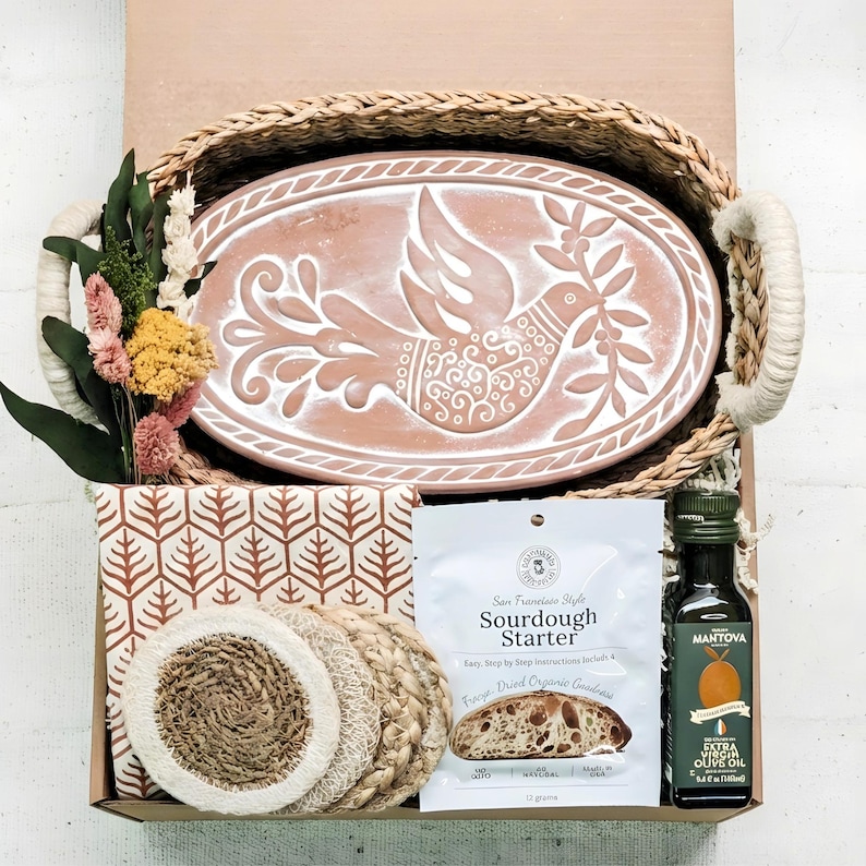 May include: A gift basket containing an oval terracotta tray with a white dove design, a patterned cloth, a packet of San Francisco Style Sourdough Starter, a bottle of Mantova extra virgin olive oil, woven coasters, and dried flowers.