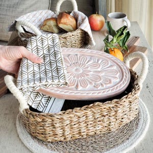 May include: A rectangular woven seagrass basket with thick rope handles, containing a light pink oval wooden lid with a carved floral motif. A white cloth napkin with a gold geometric tree pattern is draped over the basket and lid.