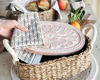 May include: A rectangular woven seagrass basket with thick rope handles, containing a light pink oval wooden lid with a carved floral motif. A white cloth napkin with a gold geometric tree pattern is draped over the basket and lid.