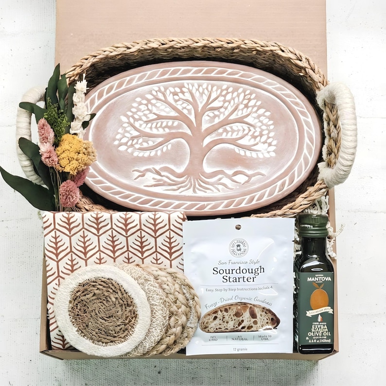 Top-down view of an artisan sourdough baking gift box from KORISSA, featuring a oval terracotta bread warmer, a handwoven seagrass basket, sourdough starter, premium olive oil, a tea towel and coasters.