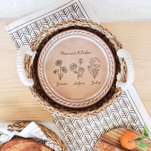 May include: A round terracotta plate with a white rope-textured rim, featuring the engraved text Mama's Kitchen, three floral illustrations, and the names James, Sophia, and Julie, nestled inside a round woven seagrass basket with white handles.
