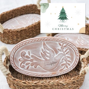 May include: Two oval-shaped ceramic trays with a white dove design and the text "Joy Miller" are displayed in woven baskets. The baskets have a natural brown color. A Christmas card with a green Christmas tree and the text "Merry Christmas" is in the background.