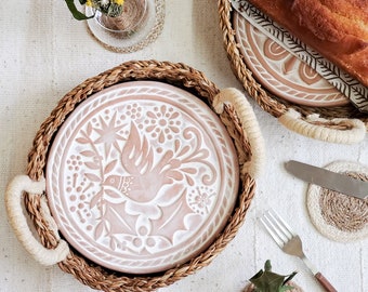 May include: Two round woven seagrass trays with cream-colored rope handles, each containing a circular ceramic plate featuring a relief-carved design of a bird, holly leaves, and floral motifs in soft terracotta and white tones.