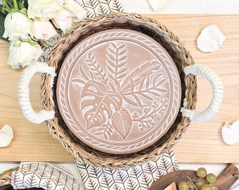 May include: A round terracotta tray with a white embossed botanical leaf pattern, featuring a woven seagrass basket rim and two thick white rope handles. The tray is circular with a flat base and a textured, natural finish on the woven border.
