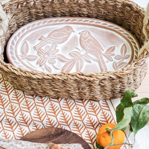 May include: A white ceramic oval serving dish with a raised design of two birds perched on branches with a flower. The dish is sitting inside a woven wicker basket.